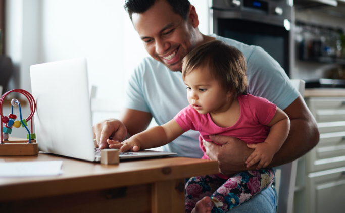 A man holding a toddler in his lap while working on a laptop computer